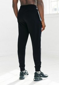 Schwarze sportliche Jogginghose mit schmaler Passform, elastischem Bund, Seitentaschen und gerippten Bündchen, kombiniert mit schwarzen Turnschuhen und grauen Socken.