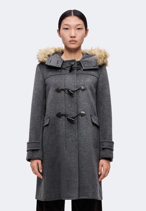 Jeune femme aux cheveux raides porte un duffle-coat gris avec une capuche bordée de fourrure et trois fermetures à boutons noirs.