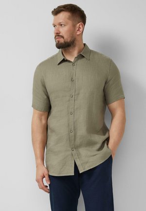 Mann mit Bart, der ein olivgrünes kurzärmeliges Hemd mit Knöpfen und marineblaue Hose trägt, steht mit einer Hand in der Tasche und schaut nach links.