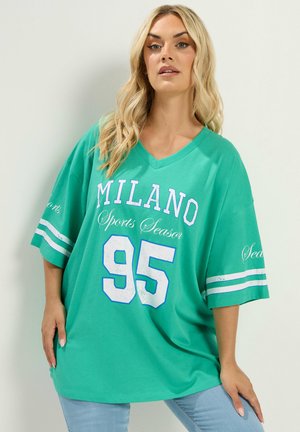 Lyshåret kvinde iført en løs grøn sportstrøje med "MILANO Sports Season 95" trykt på, poserer mod en ensfarvet lys baggrund.