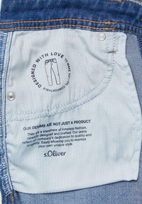 Modra denim oznaka z svetlo modrim tekstilom v ozadju. Tiskano besedilo vključuje načrte za oblikovanje in blagovno znamko "s.Oliver." Jasno vidne šivarske podrobnosti.