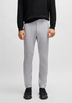 Homme portant un pantalon slim gris clair, un haut à manches longues noir et des baskets noires, debout devant un fond clair uni.