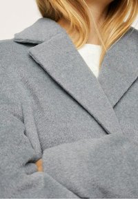 Šedý kabát z vlněné směsi s klopovým límcem a měkkým, strukturovaným povrchem. Má široké rukávy a uvolněný siluetu.