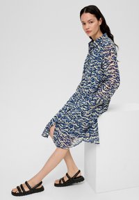 Robe à manches longues, bleue et beige à motifs, avec un devant à bouton et des poches latérales ; associée à des sandales à lanières noires.