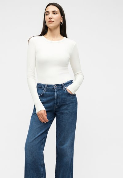 Γυναίκα με μακριά σκούρα μαλλιά που φορά ασημένια σκουλαρίκια κρίκους, λευκό εφαρμοστό τοπ με μακριά μανίκια και ψηλόμεσο φαρδύ μπλε τζιν, όρθια με το ένα χέρι στην τσέπη.