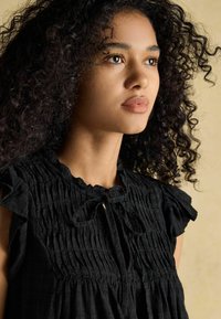 Mustast smock-kraega pluus lühikeste volangvarrukatega ja kaela juures paelaga kinnitusega, millel on tekstureeritud kangas ja lõdvema istuvusega.