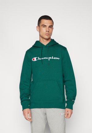Grön fleecehoodie med en frontficka, dragsko i huvan och broderad "Champion"-logotyp i vitt på bröstet. Grå sweatpants bärs nedanför.