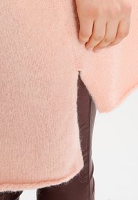 Light pink knitted sweater with a textured finish, featuring a side slit. A hand is holding the edge, revealing soft fabric details.