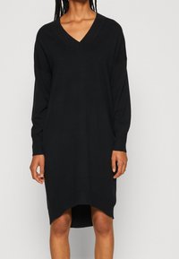 Abito nero lavorato a maglia con scollo a V profondo, maniche lunghe, polsini a coste e orlo asimmetrico. Tessuto morbido, materiale leggero per il massimo comfort.