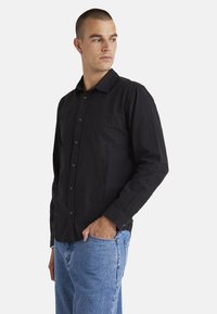 Black button-up shirt with long sleeves, a collar, and a relaxed fit, made from a textured fabric. Worn with light blue jeans.