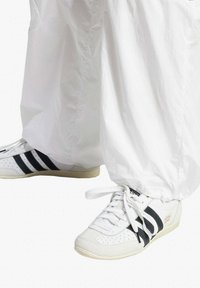 White sneakers with black stripes, perforated toe, and subtle logo, paired with white, lightweight, loose-fitting pants.