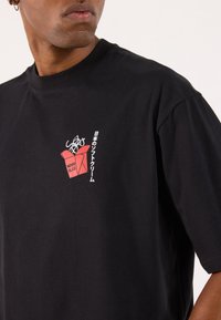 Sort bomulds t-shirt med et lille rødt grafik af en isboks med japansk tekst, korte ærmer og rund halsudskæring.