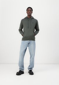 Haut à capuche vert olive avec cordon de serrage, poche kangourou, associé à un jean bleu clair à coupe large et des chaussures noires à semelle épaisse.