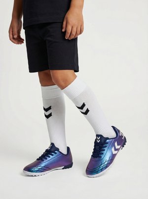 Barn iført sorte shorts, hvide knæstrømper med sorte vinkler og blå-lilla gradient fodboldstøvler med hvide vinkler.