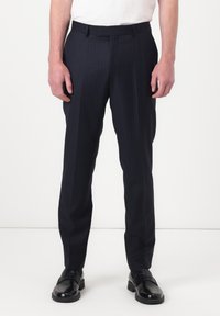 Homme portant un pantalon habillé noir à fines rayures et des mocassins en cuir noir, debout devant un fond blanc uni.