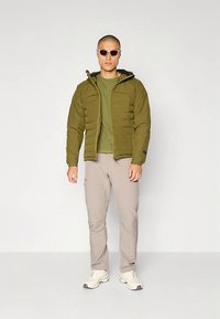 Riečiama striukė alyvuogių žalios spalvos su sagtimi priekyje ir kapišonu, derinama su žaliu marškinėliu ir šviesiai smėlio spalvos kelnėmis. Balti sportiniai bateliai ir akiniai nuo saulės.