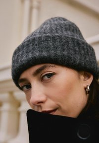 Berretto in maglia grigio con texture a coste, indossato leggermente inclinato. Primo piano sul volto di una persona, che indossa un cappotto nero e orecchini a cerchio argento.