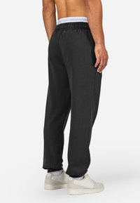 Man wearing black elastic-waist sweatpants, white socks, and white sneakers, standing with back facing the camera on a white background.