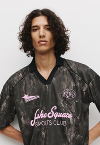 Sort trøje med en marmoreringstekstur, udstyret med en sort krave, lyserød skrift for "New Heights" og "Jha Square Sports Club" samt et rundt logo.