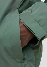 Grønn vanntett jakke med brun innerfòr, med sidelomme og flate ermekanter. Jevn tekstur, minimalistisk design.