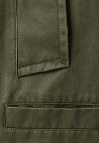 Gros plan sur un tissu en daim vert avec des coutures détaillées sur deux rabats de poche rectangulaires, montrant la texture et les lignes de couture.
