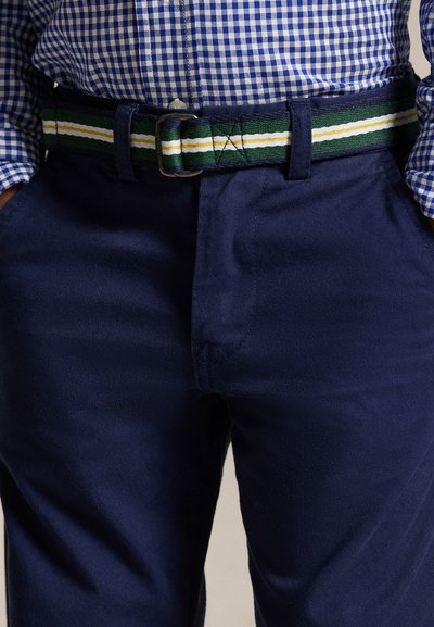 Persona que lleva pantalones azul marino con un cinturón de tela a rayas en verde, blanco y amarillo y una camisa azul de cuadros metida dentro.