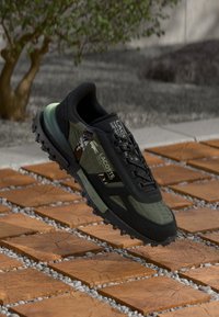 Scarpa atletica nera e verde oliva con tomaia testurizzata, chiusura con lacci, suola in gomma e accenti mimetici. Elevata sopra una superficie in pietra.