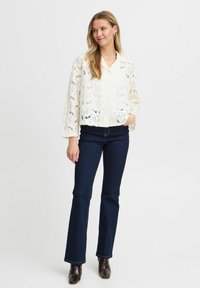 Blus i spets i off-white med blommönster, långa ärmar, knäppning framtill, ihop med mörkblå utsvängda jeans och bruna ankelskor.