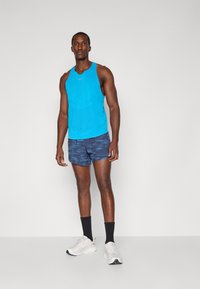 Sportoutfit mit einem leuchtend blauen ärmellosen Oberteil und dunkelblauen gemusterten Shorts, kombiniert mit weißen Laufschuhen und schwarzen Socken.