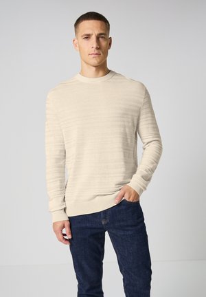 Maglione beige lavorato a maglia con righe testurizzate, scollatura rotonda e polsini a coste. Abbinato a jeans in denim blu scuro. Design sottile e discreto.