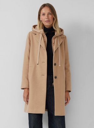 Manteau beige au tissu doux, avec capuche et boutons-pression. Il possède deux poches latérales et un col. Porté par-dessus un pull à col roulé noir.