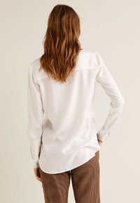 Camicia bianca a maniche lunghe con orlo curvo, texture liscia e polsini con bottoni. La vista posteriore mostra dettagli cuciti e un design leggermente sagomato.