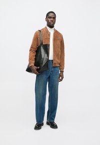 Rjava semiš jakna, bel puli, modre široke jeans hlače in črna usnjena torbica. Model nosi črne čevlje, stoji ob preprostem ozadju.