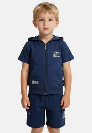 Jeune garçon aux cheveux blonds portant un sweat à capuche zippé bleu marine à manches courtes et un short assorti avec des logos de marque.