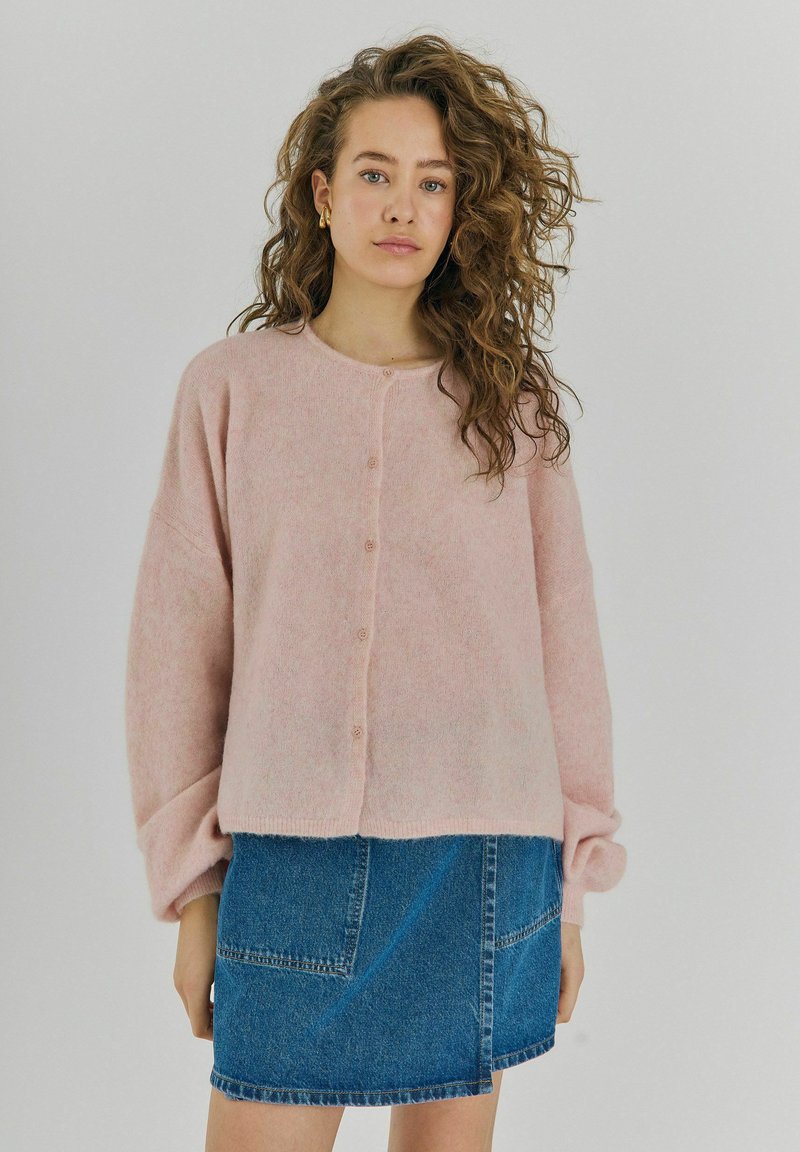 Světle růžová pletená kardigán se zaobleným výstřihem, předním zapínáním na knoflíky a oversized rukávy. V kombinaci s modrou džínovou sukní.