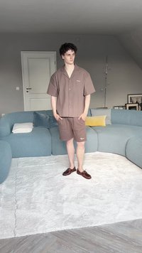 Barna, rövid ujjú ing és hozzá illő, könnyű anyagból készült rövidnadrág. Barna cipő. Egy modern nappaliban áll, ahol egy ívelt kanapé található.