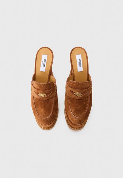 Mules slip-on coklat suede dengan detail perangkat keras warna emas dan insole kulit coklat yang bertuliskan "Moschino Milano" di latar belakang putih.