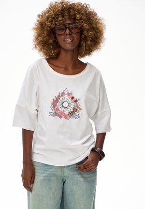 Kobieta o kręconych włosach, nosząca duże okulary przeciwsłoneczne, kolczyki koła, biały t-shirt z kwiatowym nadrukiem, jasnoniebieskie dżinsy i ciemną bransoletkę.