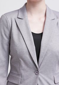 Jasnoszary żakiet z teksturowanej tkaniny, z klapami w kształcie notcha, podwójnymi przednimi kieszeniami i pojedynczym zapięciem na guzik. Noszony na czarnym topie.