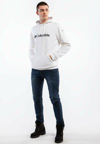Biała bluza z kapturem z logo "Columbia", z przednią kieszenią i sznurkiem w kapturze. W zestawieniu z ciemnymi dżinsami i czarnymi sznurowanymi butami.