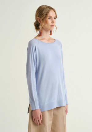 Frau mit blondem Haar in einem niedrigen Dutt, die einen hellblauen Pullover mit langen Ärmeln und beige Hosen trägt und auf einem schlichten Hintergrund nach rechts schaut.