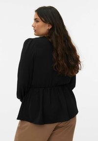 Blouse noire avec un ourlet à basque, tissu texturé, manches bouffantes et col rond, associée à un pantalon beige.