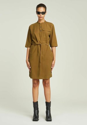 Krótkosleeve oliwkowa sukienka z kołnierzykiem, zapięciem na guziki, paskiem w talii i kieszonką z przodu. Modelka nosi czarne okulary przeciwsłoneczne i czarne kozaki do kolan.