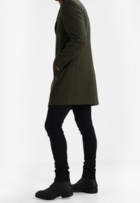 Manteau long vert olive à la coupe cintrée avec de discrètes épaulettes, associé à un jean noir taille fine et des bottes noires à lacets.