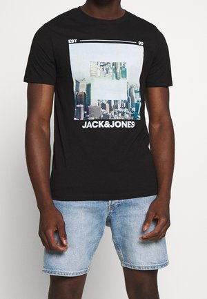 Mężczyzna noszący czarną koszulkę z grafiką panoramy miasta i tekstem "JACK & JONES", w połączeniu z jasnoniebieskimi szortami jeansowymi.
