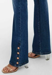 Pieds portant des talons ouverts transparents à talons carrés, et un jean en denim bleu foncé avec quatre boutons en cuivre le long de la couture extérieure de la cheville.