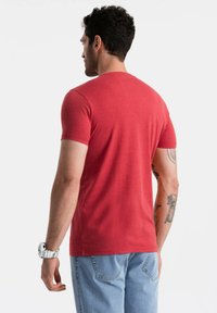 Červené tričko s krátkým rukávem vyrobené z měkké látky. Má klasický kulatý výstřih a slim fit střih. Nosí se s světle modrými džíny a bílými hodinkami.