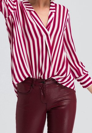 Vrouw draagt een wit overhemd met lange mouwen met verticale rode strepen en glanzende donkerrode broek met hoge taille.