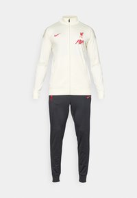 LFC Nike trenirka koja se sastoji od kremaste jakne sa zipom i crnih hlača, obe sa crvenim logotipima. Glatka tkanina, uski kroj i rebrasti manžeti.
