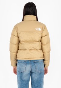 Beige gewatteerde jas met hoge kraag en logo op de achterkant. Bevat horizontale naden en ribbelmanchetten. Gekozen in combinatie met blauwe denim jeans.